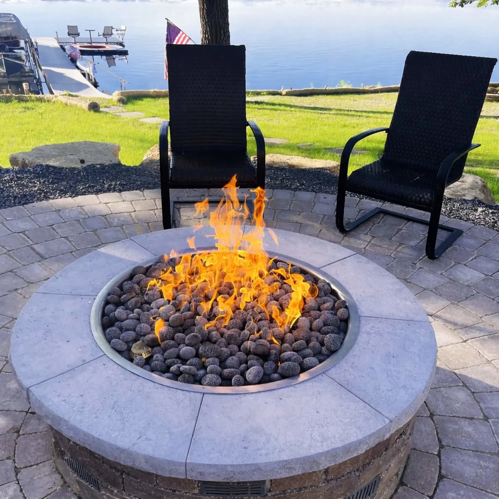 Evening backyard scene featuring a round stone fire pit table with active flames illuminating surrounding Adirondack chairs. The fire pit is integrated into a raised stone patio with built-in seating, creating a warm and inviting outdoor gathering space under a deep blue twilight sky.