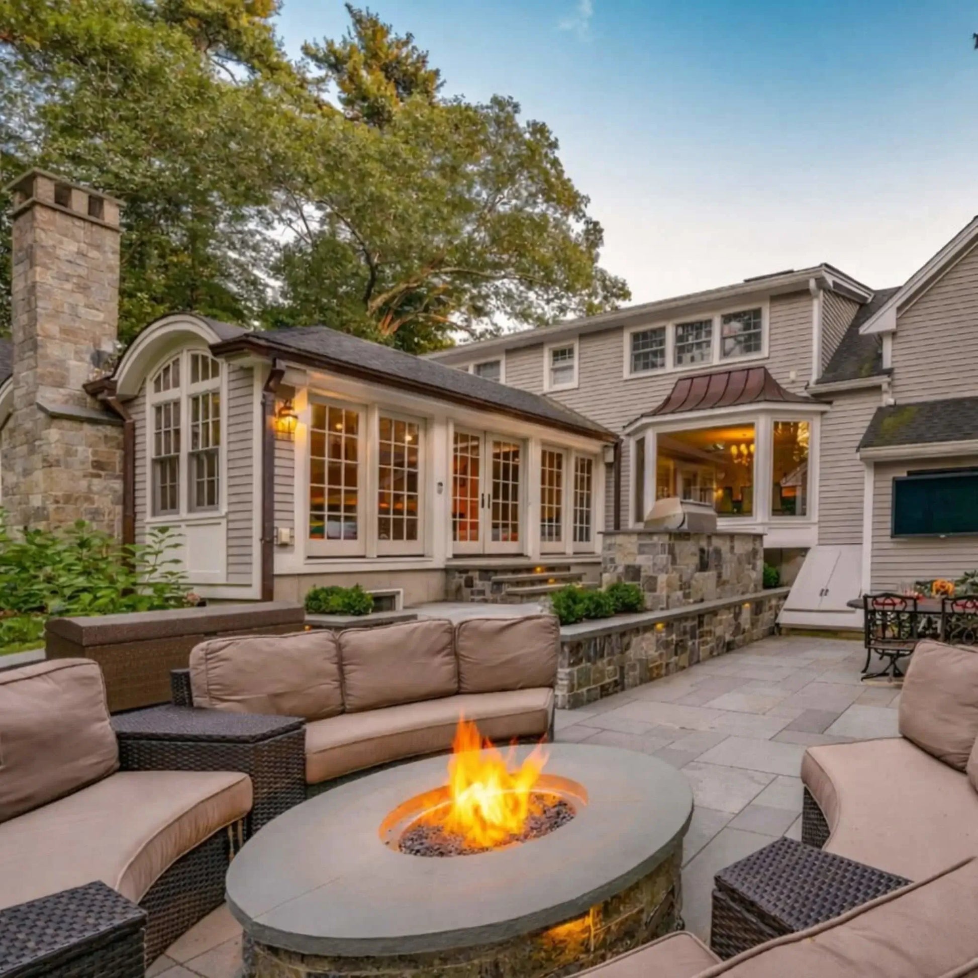 Lakeside patio with a circular gas fire pit table filled with lava rock and surrounded by two black outdoor chairs. Bright flames rise in the center while a dock, calm water, and distant shoreline create a relaxing waterfront setting ideal for outdoor lounging.