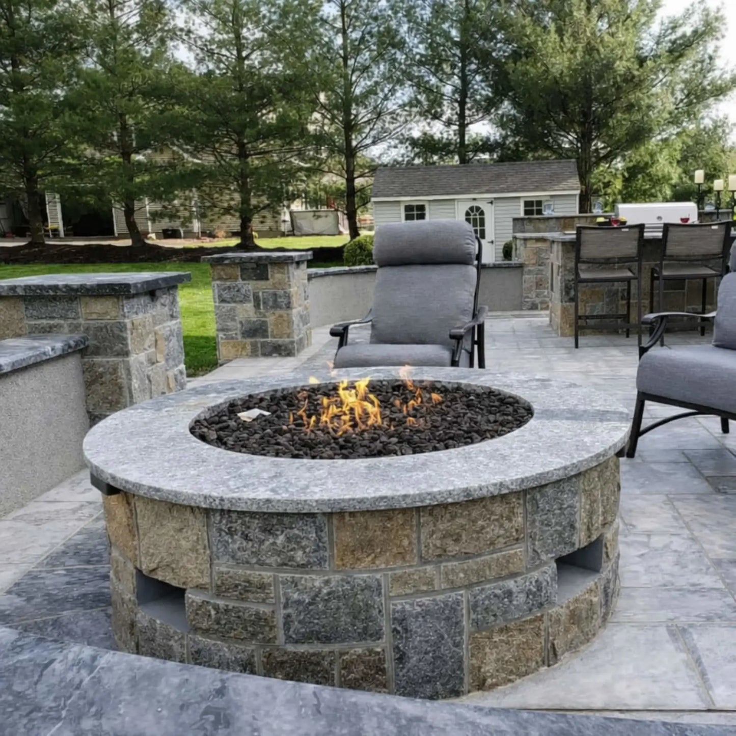 Elegant backyard patio featuring a round fire pit table with a smooth top and glowing flame centerpiece, surrounded by wicker sectional seating. The space connects to a large home with expansive windows, stone accents, and warm lighting, creating a high-end outdoor living environment.