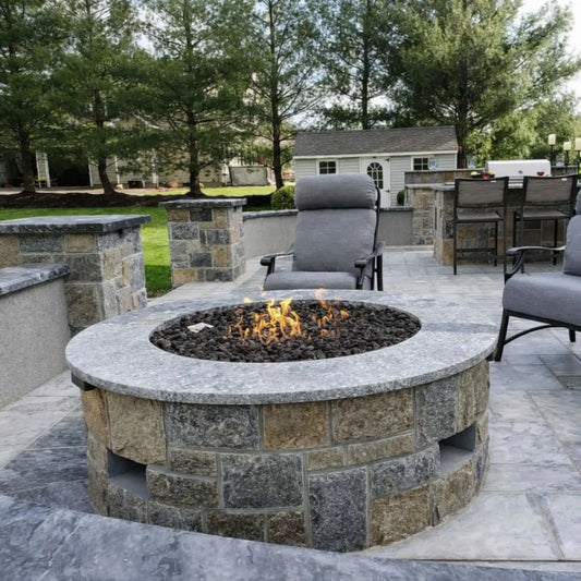 Elegant backyard patio featuring a round fire pit table with a smooth top and glowing flame centerpiece, surrounded by wicker sectional seating. The space connects to a large home with expansive windows, stone accents, and warm lighting, creating a high-end outdoor living environment.