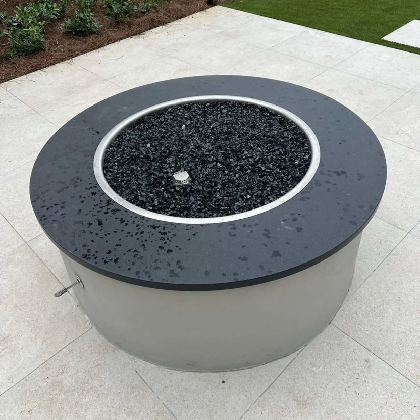 HPC Fire Inspired Hearth Products electronic ignition round drop in penta burner installed in a modern circular fire pit filled with black fire glass. The unlit burner sits centered within a sleek patio setup, surrounded by light stone pavers and clean landscaping, showcasing a contemporary outdoor fire feature design.