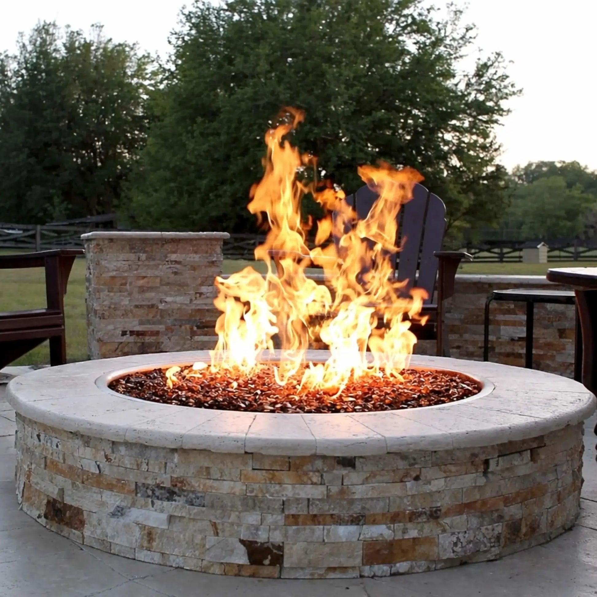 HPC Fire Inspired Hearth Products electronic ignition penta burner producing tall, vibrant flames over decorative fire glass in a stone fire pit. The backyard setting includes Adirondack chairs, stone columns, and greenery, capturing a warm, inviting outdoor entertainment space with strong flame presentation.