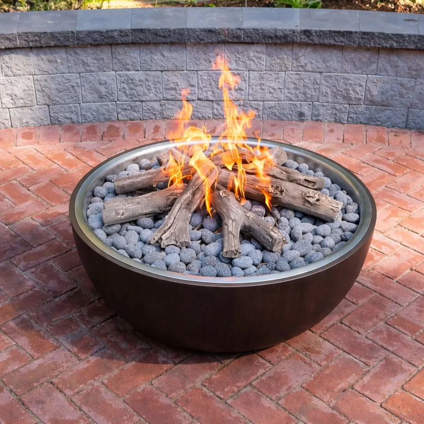 HPC Fire Inspired Hearth Products electronic ignition penta burner installed in a modern round metal fire bowl with ceramic logs over gray lava rock. Flames rise naturally through the log set, placed on a red brick patio with a stone retaining wall behind, showcasing a traditional campfire look with gas convenience.