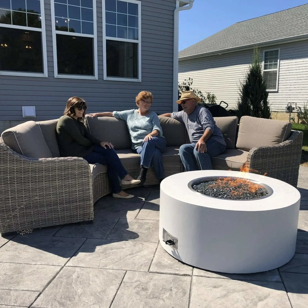 HPC Fire Inspired Hearth Products round drop in electronic ignition penta burner showcased in a residential patio scene with three people relaxing on a sectional sofa. The fire pit produces steady flames over black fire glass, highlighting comfort, social gathering, and real-life backyard use.
