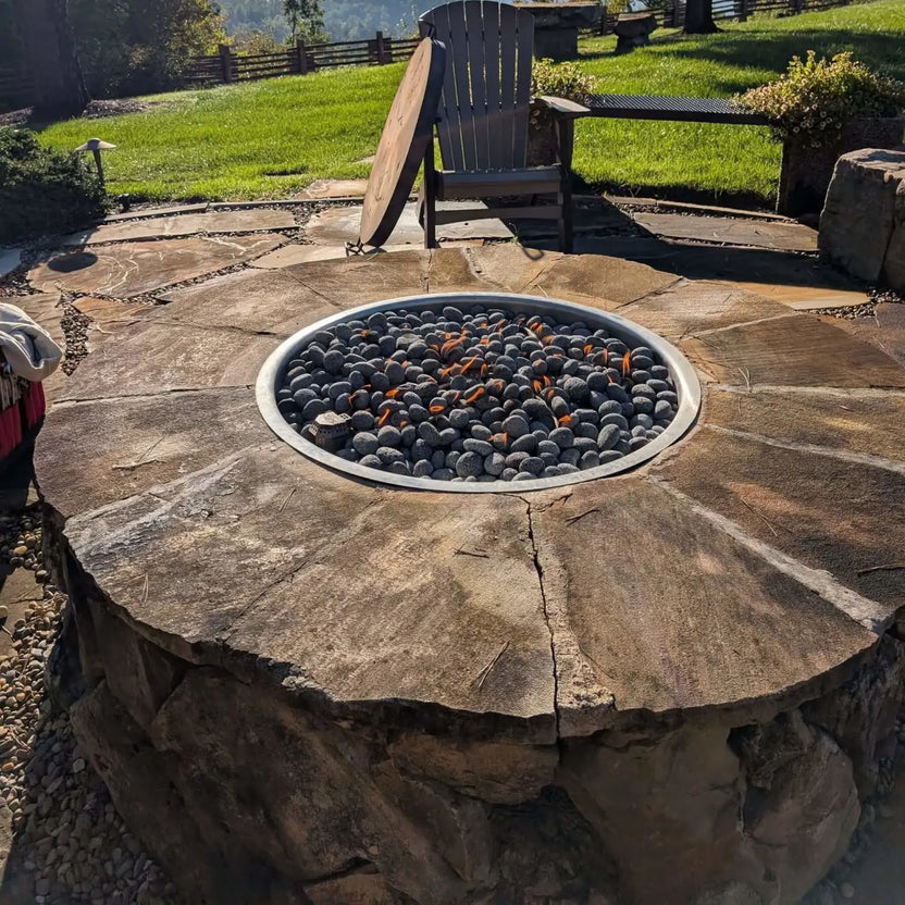 HPC Fire Inspired Hearth Products round drop in penta burner installed in a rustic stone fire pit with natural slab top. Lava rock fills the interior while small flames flicker across the surface, surrounded by a scenic outdoor setting with grass, patio furniture, and natural stone landscaping.