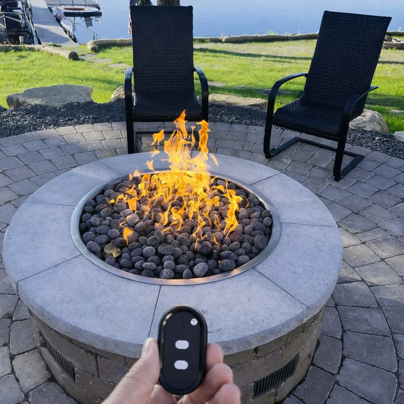 HPC Fire Inspired Hearth Products Controls Round Drop In FPPK torpedo penta burner shown in use with a handheld remote control in the foreground. The fire pit is filled with smooth lava stones, producing tall yellow flames, while two patio chairs and a waterfront view in the background emphasize convenience and remote ignition functionality.