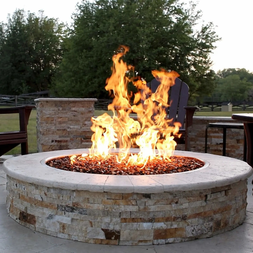 HPC Fire Inspired Hearth Products FPPK push button flame sensing torpedo penta burner featured in a modern circular stone fire pit with reflective fire glass. Strong flames rise at the center, surrounded by a well-designed patio with stone seating walls and outdoor chairs, creating a warm and inviting backyard gathering area.