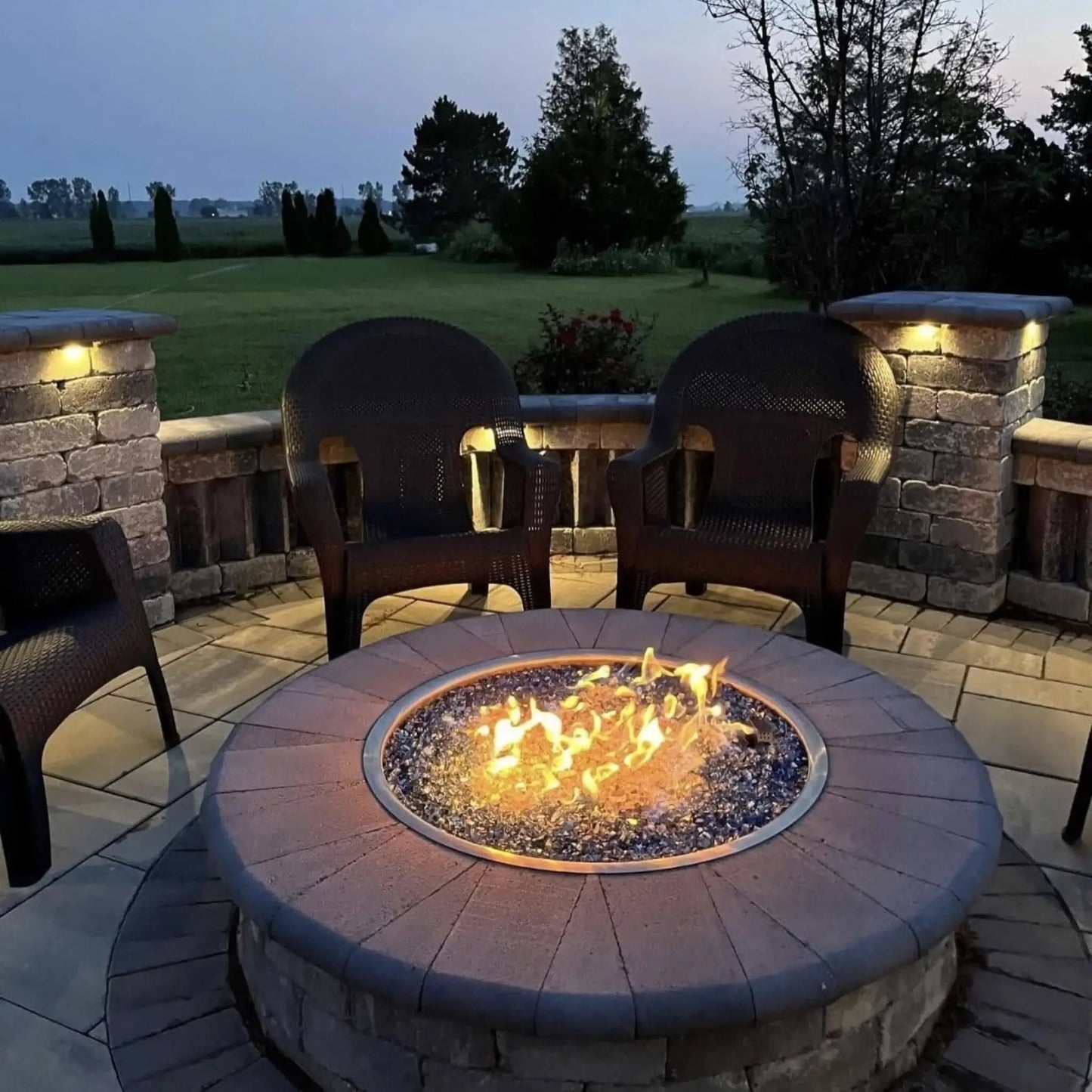 HPC Fire Inspired Hearth Products Controls Round Drop In FPPK torpedo penta burner glowing at dusk in a backyard patio. The fire pit emits warm flames over glass media, surrounded by patio chairs and stone seating walls, creating a cozy outdoor ambiance with built-in lighting and structured landscaping.