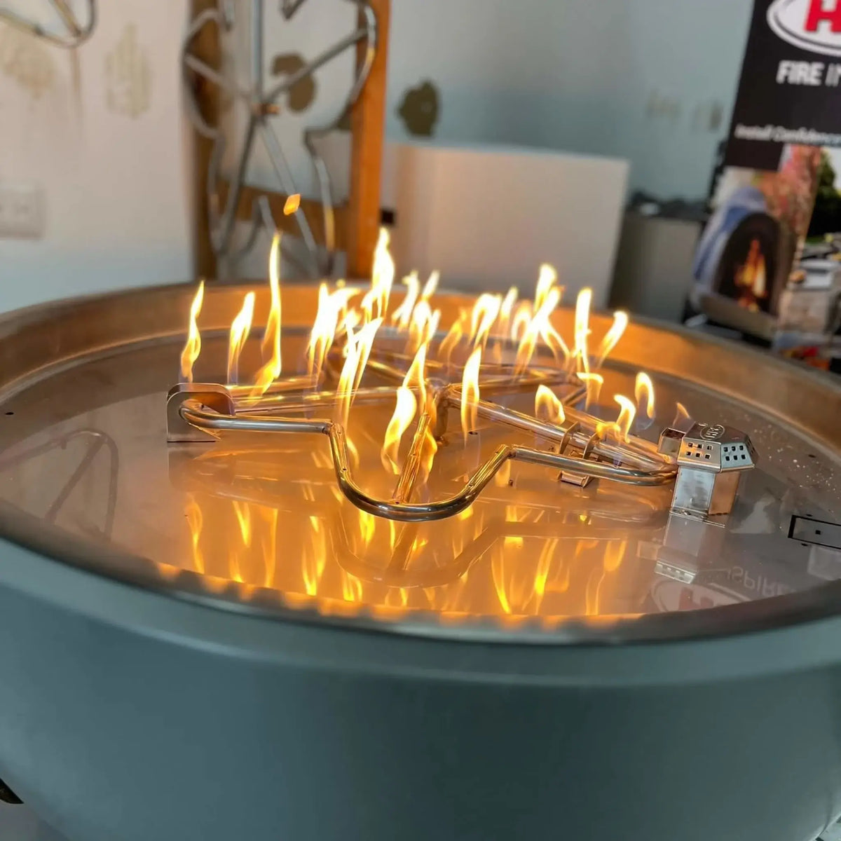HPC Fire Inspired Hearth Products FPPK push button flame sensing torpedo penta burner captured in a close-up with active flames dancing across a stainless steel burner. The reflection of fire on the metal pan emphasizes ignition performance and flame consistency within a modern fire pit enclosure.