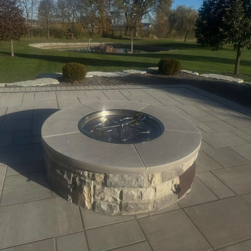 HPC Fire Inspired Hearth Products Controls Round Drop In FPPK torpedo penta burner installed in a large circular stone fire pit on a spacious paver patio. The stainless steel burner sits recessed in the center, unlit, with a scenic backyard featuring trimmed shrubs, open lawn, and a small pond in the distance, highlighting clean installation and outdoor landscape integration.