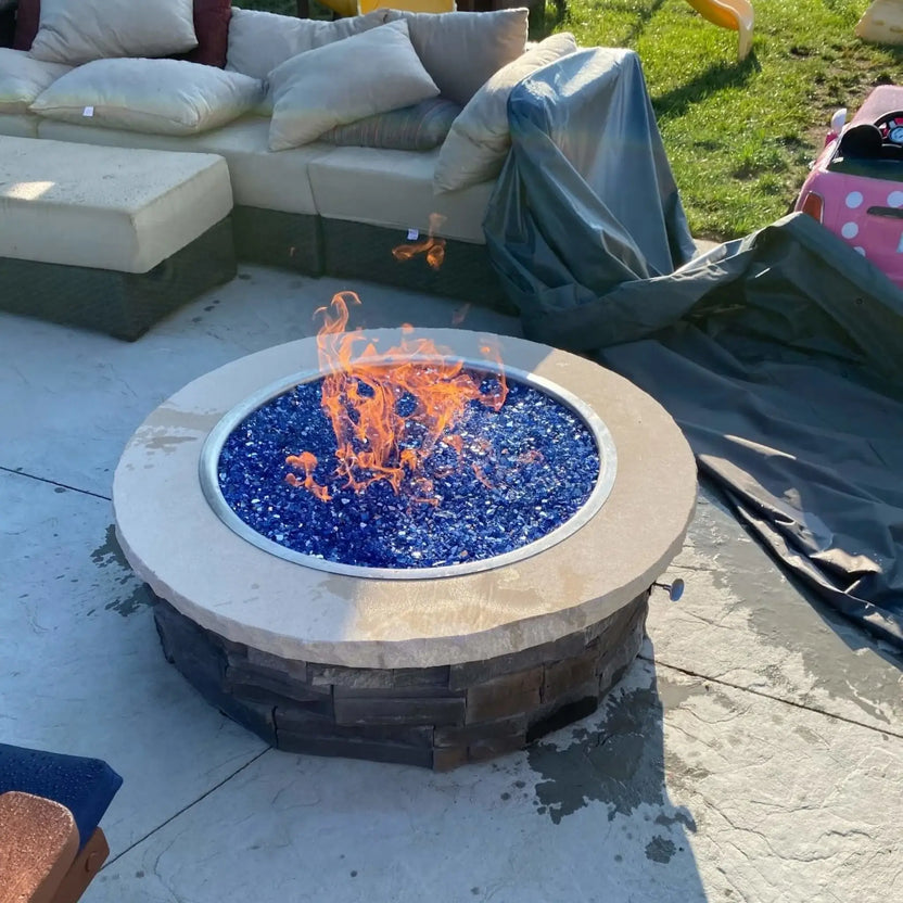 HPC Fire Inspired Hearth Products torpedo penta burner showcased in a backyard fire pit filled with vibrant blue fire glass and active flames. The round drop in match lit CSA MLFPK FLEX system is installed in a stone fire feature beside outdoor seating, emphasizing bold flame contrast and modern fire glass aesthetics.