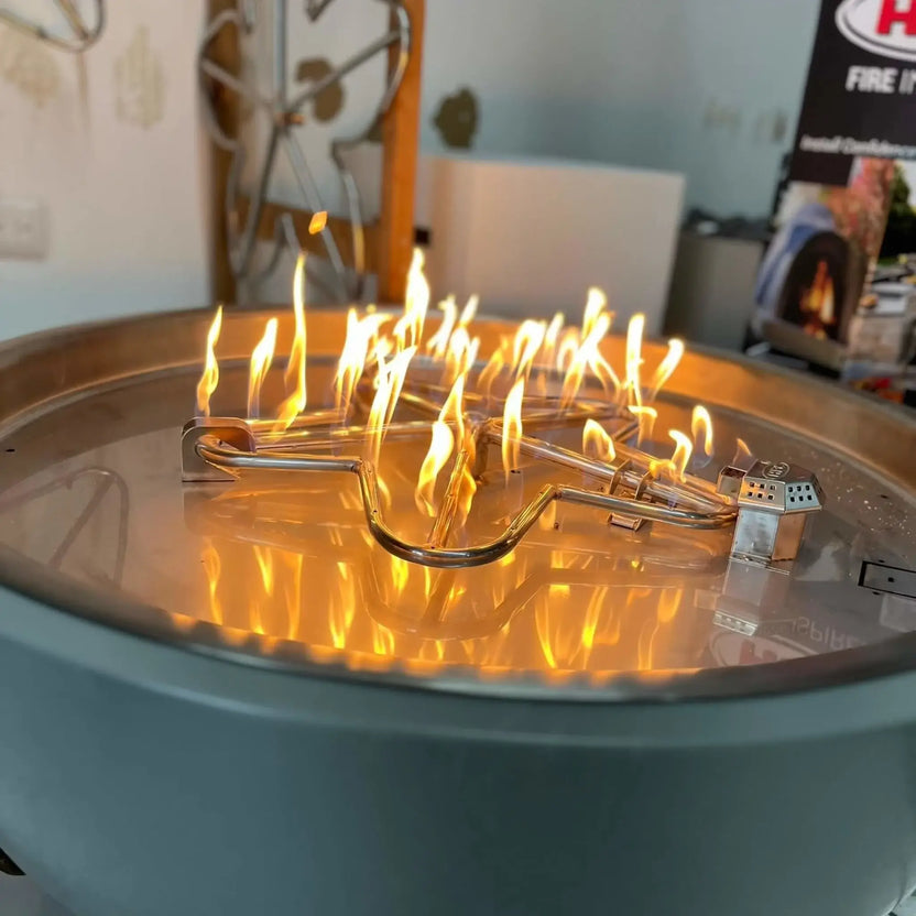 HPC Fire Inspired Hearth Products torpedo penta burner integrated into a circular fire pit with gray lava rock, producing steady yellow flames. The top-down perspective highlights the burner’s even heat distribution and the clean stainless steel drop-in pan design.