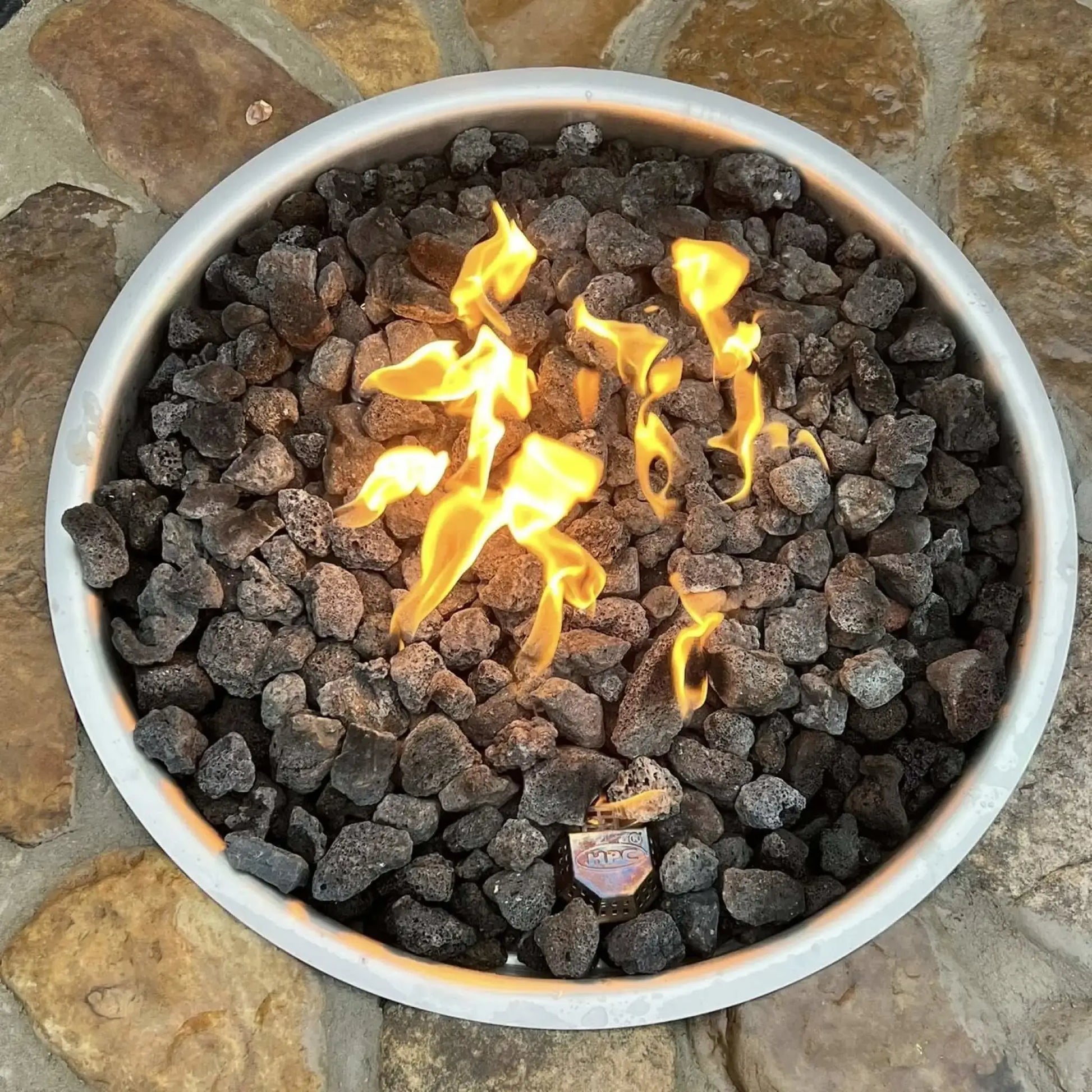 HPC Fire Inspired Hearth Products torpedo penta burner showcased in a modern outdoor fire pit with smooth lava stones and minimal flame output. The natural setting with seating and patio stonework highlights a relaxed outdoor environment using the MLFPK FLEX system.
