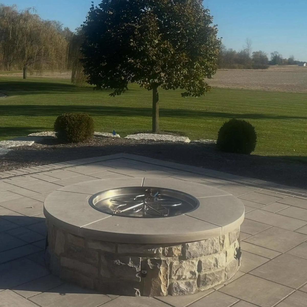 HPC Fire Inspired Hearth Products round drop in match lit torpedo penta burner installed in a circular stone fire pit on a spacious patio. The image captures the burner pan and surrounding landscape, emphasizing durable materials, clean installation, and integration into outdoor living design.