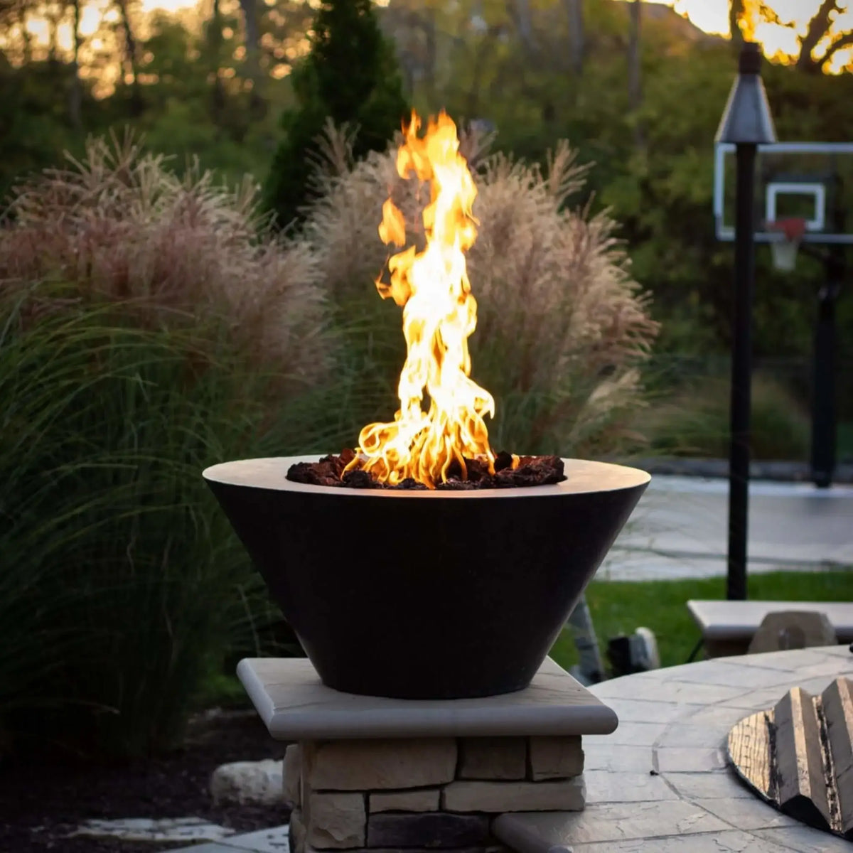 HPC Fire Inspired Hearth Products round flat electronic ignition on off hi lo low standard torpedo penta burner showcased in a modern outdoor fire bowl on a stone pedestal. Bright flames rise over lava rock with ornamental grasses and a landscaped backyard behind, highlighting a clean, upscale fire feature installation at dusk.