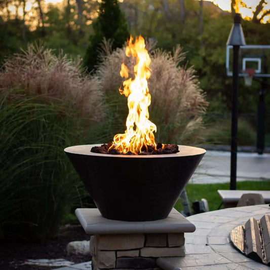 HPC Fire Inspired Hearth Products round flat electronic ignition on off hi lo low standard torpedo penta burner showcased in a modern outdoor fire bowl on a stone pedestal. Bright flames rise over lava rock with ornamental grasses and a landscaped backyard behind, highlighting a clean, upscale fire feature installation at dusk.