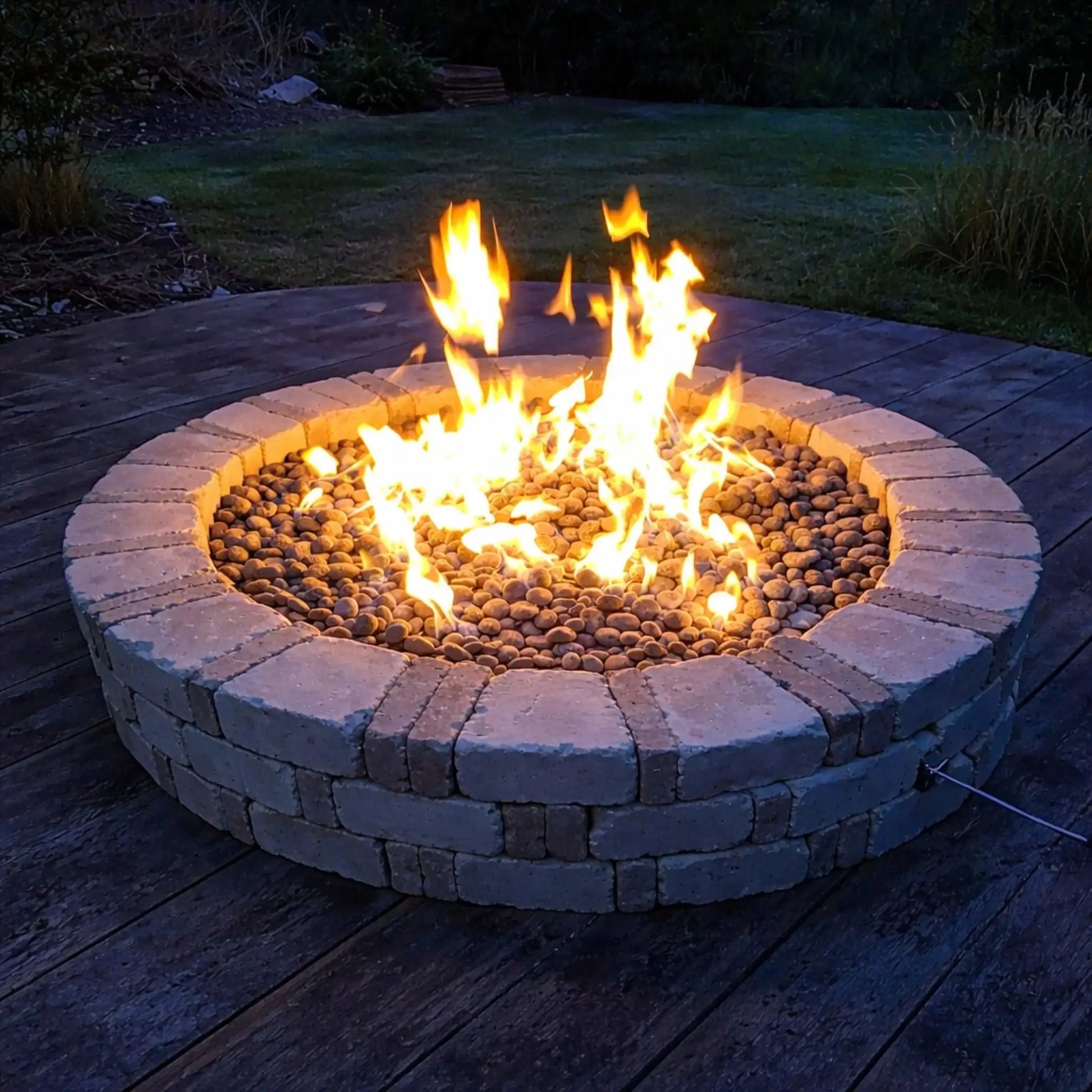 HPC Fire Inspired Hearth Products round flat electronic ignition on off hi lo low standard torpedo penta burner burning brightly in a low-profile circular fire pit on a wooden deck. Smooth river rock surrounds the flame, creating a warm glow during dusk, with a simple backyard lawn setting that emphasizes cozy outdoor relaxation.