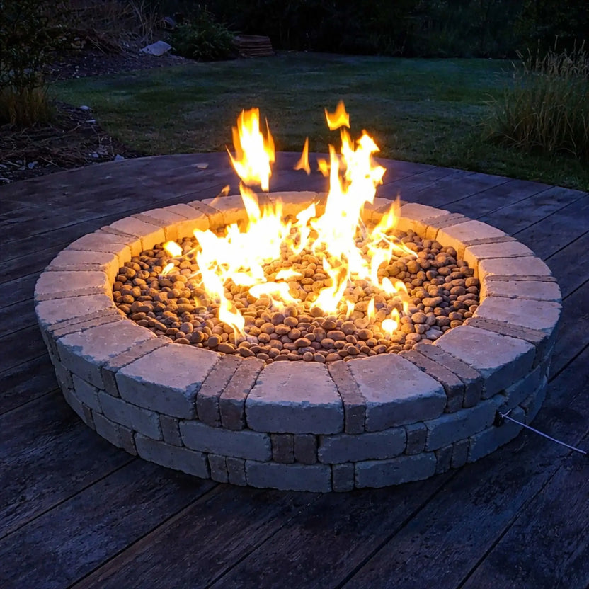 HPC Fire Inspired Hearth Products round flat electronic ignition on off hi lo low standard torpedo penta burner burning brightly in a low-profile circular fire pit on a wooden deck. Smooth river rock surrounds the flame, creating a warm glow during dusk, with a simple backyard lawn setting that emphasizes cozy outdoor relaxation.