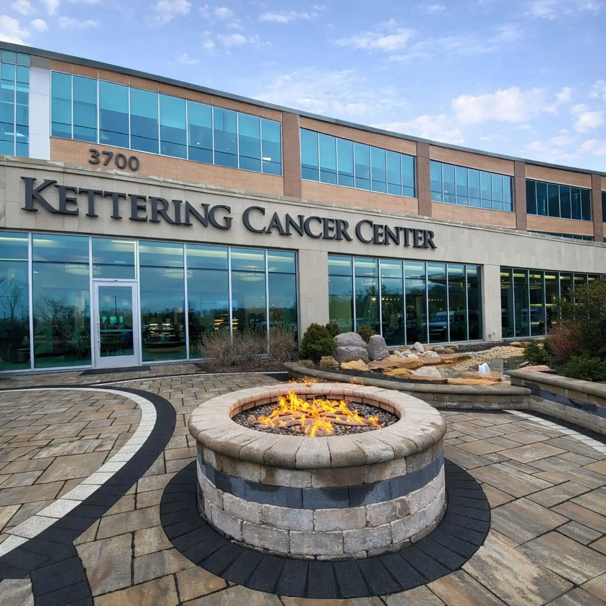 HPC Fire Inspired Hearth Products round flat electronic ignition on off hi lo low standard torpedo penta burner placed in front of a commercial building labeled Kettering Cancer Center. The circular fire pit sits within a modern paved courtyard, demonstrating a professional outdoor installation in a public or commercial landscape.