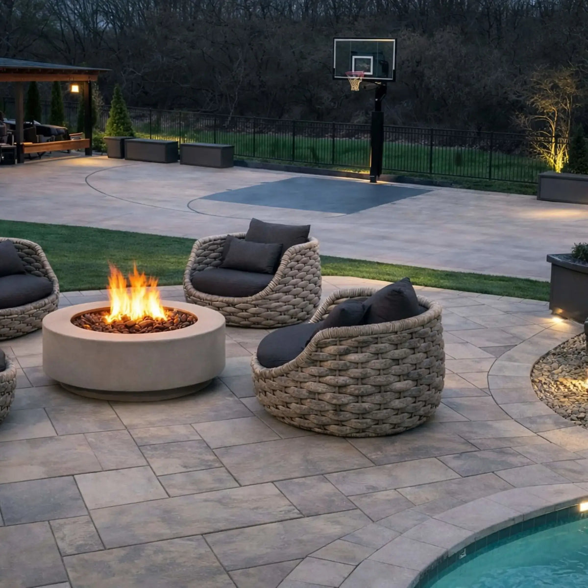 HPC Fire Inspired Hearth Products round flat electronic ignition on off hi lo low standard torpedo penta burner displayed in a luxury backyard patio with woven lounge chairs and a circular fire pit. Overlooking a basketball court and landscaped yard, the scene captures a high-end outdoor entertainment space at dusk.