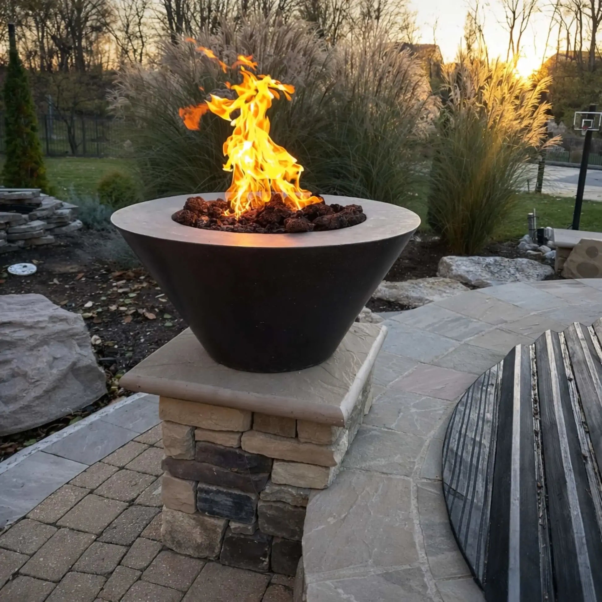 HPC Fire Inspired Hearth Products round flat electronic ignition on off hi lo low standard torpedo penta burner featured in a modern bowl-style fire feature mounted on a stone pillar. Flames rise above lava rock at sunset, surrounded by ornamental grasses and a landscaped patio, creating a warm, upscale backyard focal point.