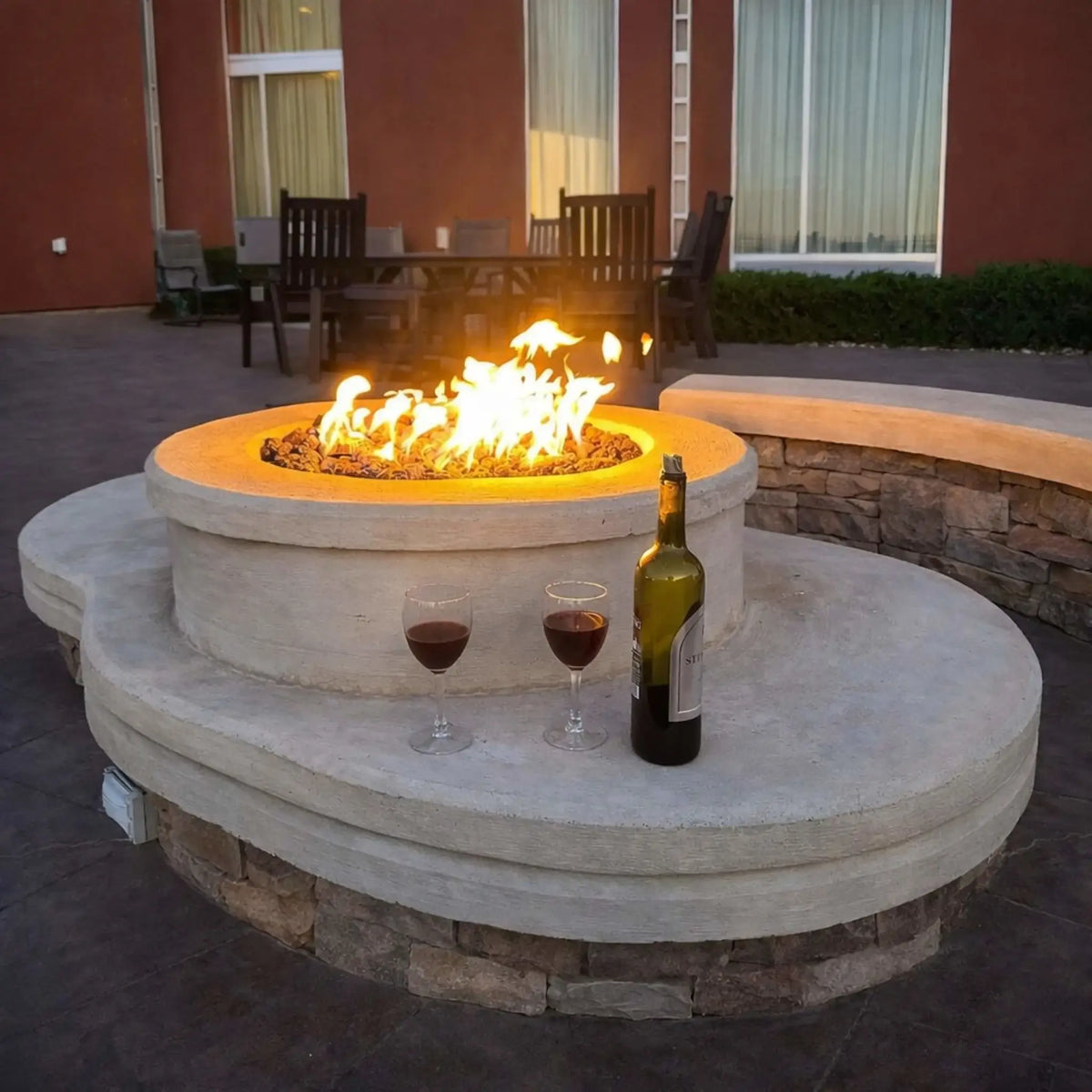 HPC Fire Inspired Hearth Products round flat electronic ignition on off hi lo low standard torpedo penta burner integrated into a modern concrete fire pit table. Flames glow warmly while a wine bottle and glasses sit nearby, set against a residential patio with outdoor dining furniture, highlighting a cozy evening entertainment setup.