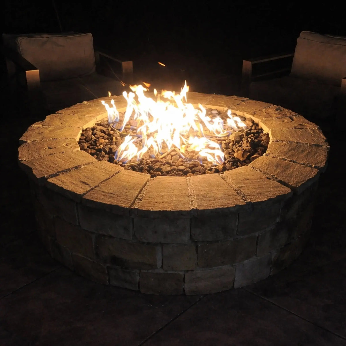 HPC Fire Inspired Hearth Products round flat electronic ignition on off hi lo low standard torpedo penta burner burning brightly in a circular stone fire pit at night. Surrounded by cushioned outdoor chairs, the fire reflects off dark stone surfaces, creating a warm and inviting nighttime patio atmosphere.