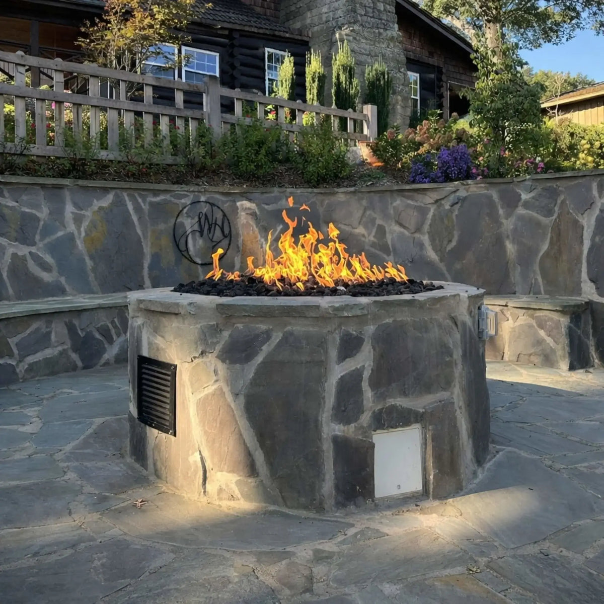 HPC Fire Inspired Hearth Products round flat electronic ignition on off hi lo low standard torpedo penta burner installed in a stone-clad fire pit with built-in lighting. Flames rise above black lava rock with a rustic cabin and landscaped retaining wall behind, highlighting a high-end residential fire feature installation.