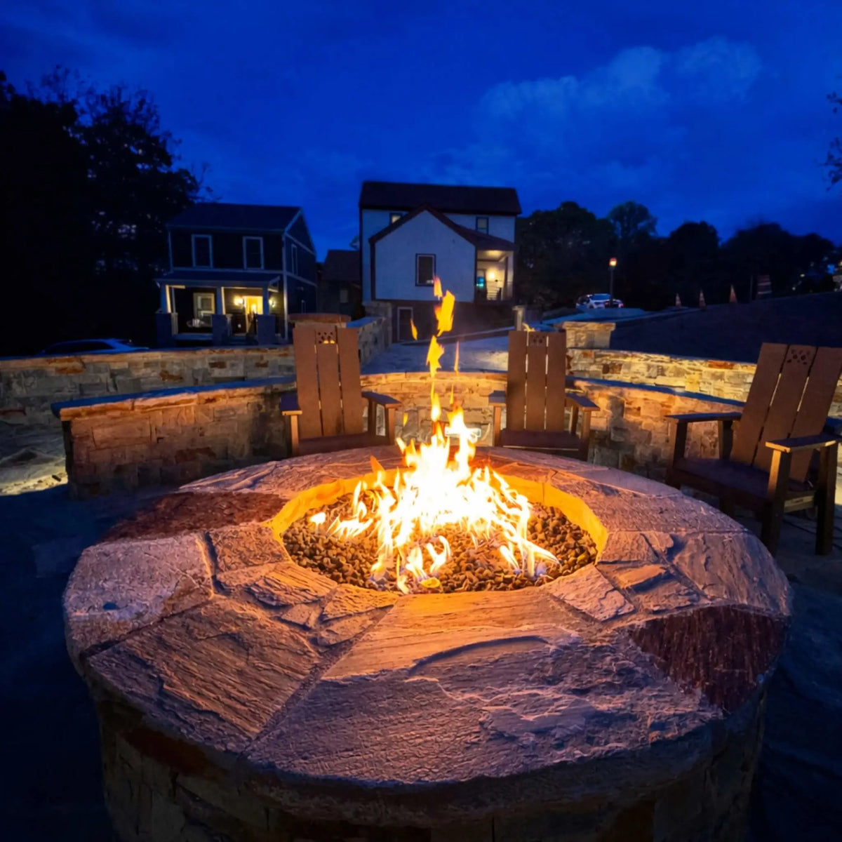 HPC Fire Inspired Hearth Products round flat FPPK push button flame sensing standard torpedo penta burner showcased in a circular stone fire pit at dusk. Bright flames rise from lava rock media, surrounded by Adirondack chairs and a curved stone seating wall, with residential homes softly lit in the background, creating a warm and inviting backyard gathering space.