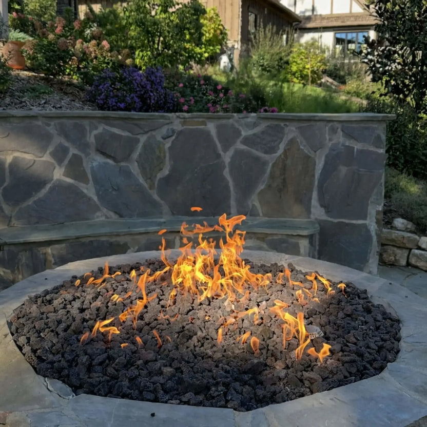 HPC Fire Inspired Hearth Products round flat FPPK push button flame sensing standard torpedo penta burner shown in a close-up of a circular fire pit filled with black lava rock. Flames spread evenly across the burner, with a stone retaining wall and garden plants in the background, emphasizing consistent flame pattern and burner performance.