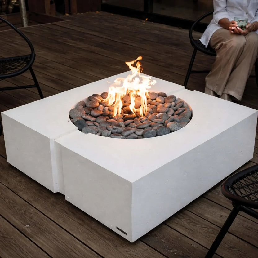 HPC Fire Inspired Hearth Products round flat FPPK push button flame sensing standard torpedo penta burner installed in a modern square white concrete fire table. Flames rise from a bed of smooth river rocks, placed on a wooden deck with outdoor seating, showcasing a contemporary fire feature design ideal for patios and entertaining.