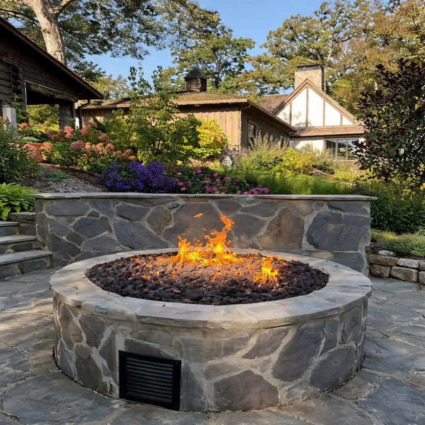 HPC Fire Inspired Hearth Products round flat match lit CSA MLFPK FLEX standard torpedo penta burner displayed in a beautifully landscaped backyard with stone retaining walls and colorful garden beds. The fire pit sits at the center with soft flames over lava rock, blending seamlessly into a well-designed outdoor space.