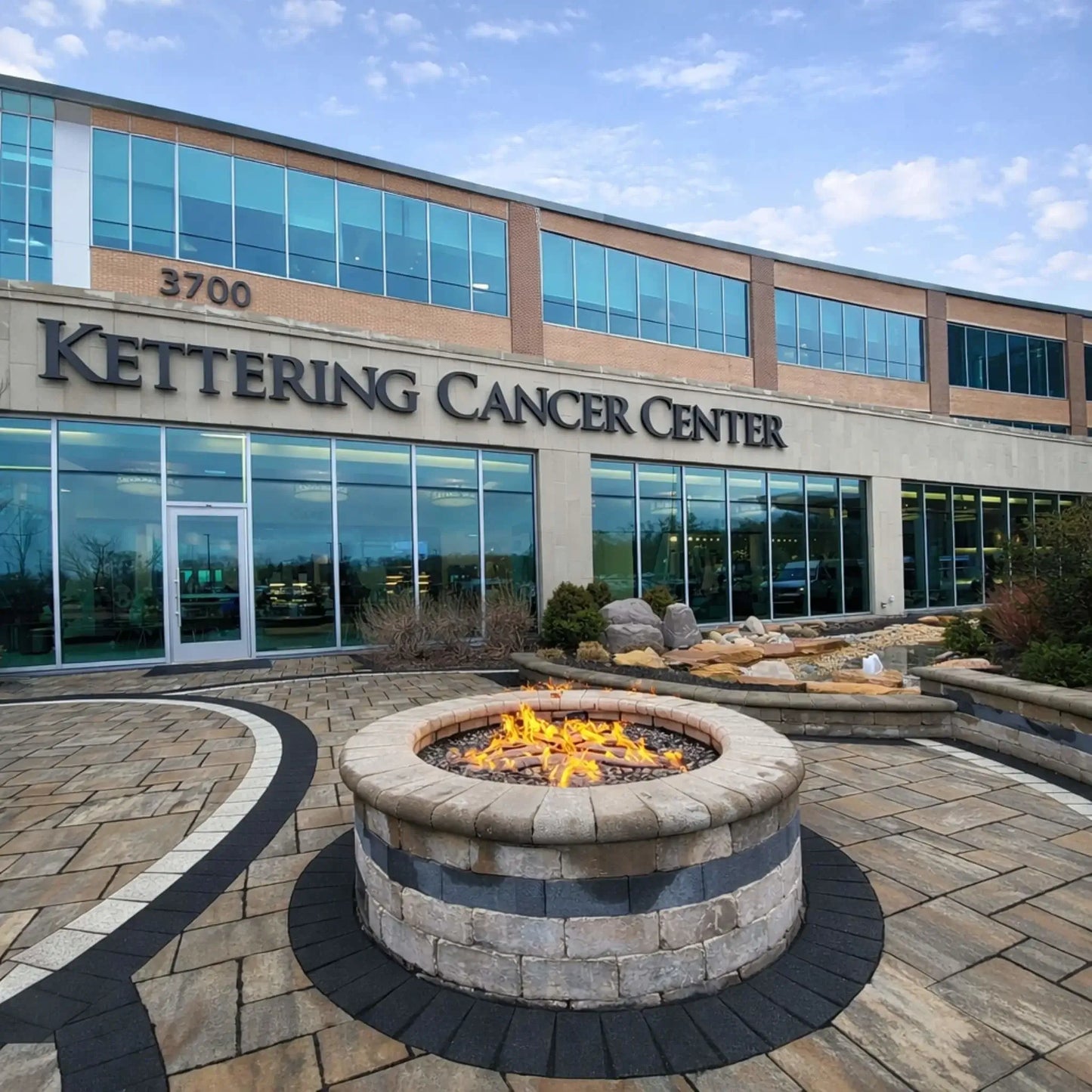 HPC Fire Inspired Hearth Products round flat match lit CSA MLFPK FLEX standard torpedo penta burner installed in a commercial-style outdoor patio in front of the Kettering Cancer Center building. A circular fire pit with clean stonework and active flames creates a welcoming, modern entrance feature.