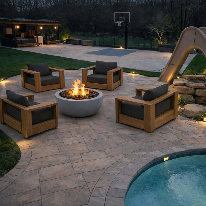 HPC Fire Inspired Hearth Products round flat match lit CSA MLFPK FLEX standard torpedo penta burner installed in a contemporary backyard patio with four wooden lounge chairs arranged around a circular fire pit. Soft lighting, a nearby pool, and a sleek slide create a modern outdoor retreat, with flames providing a warm focal point for evening gatherings.