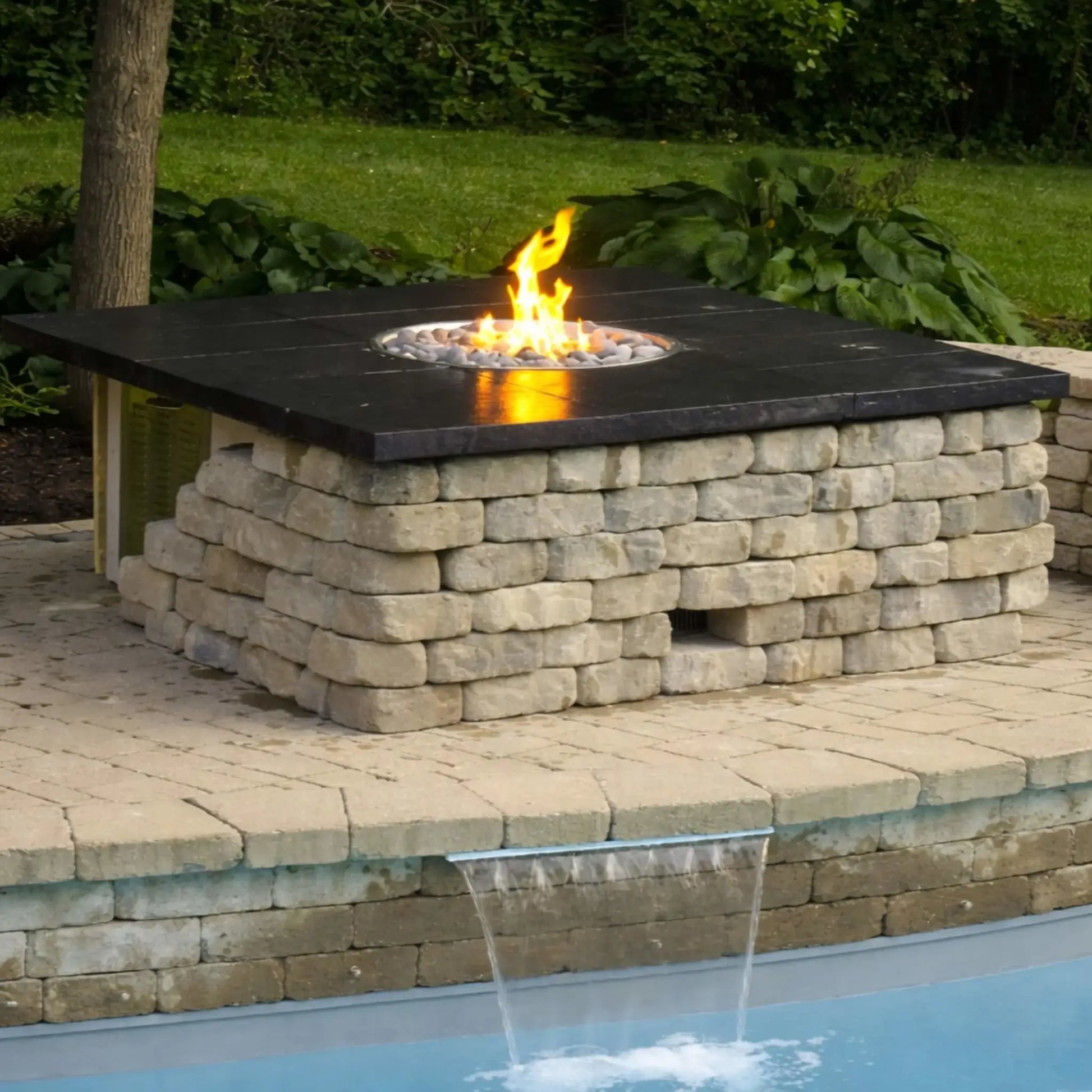 Finished square fire pit table with a dark stone top and stacked block base, centered with a round gas burner producing a bright flame over decorative fire media. Installed beside a pool with a water feature, this setup highlights a modern outdoor living space with integrated fire and water elements.