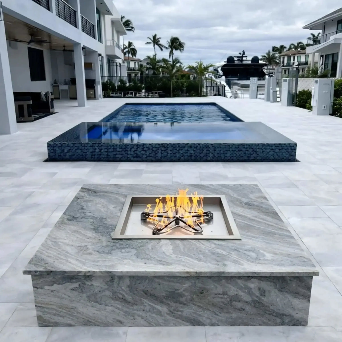HPC Fire Inspired Hearth Products square drop-in electronic ignition on off hi lo low standard torpedo penta burner installed in a sleek waterfront patio, featuring a gray stone fire table with a stainless steel burner and bright flames. The background includes a modern pool, docked yacht, canal waterway, and upscale homes, showcasing a luxurious outdoor fire feature setup.