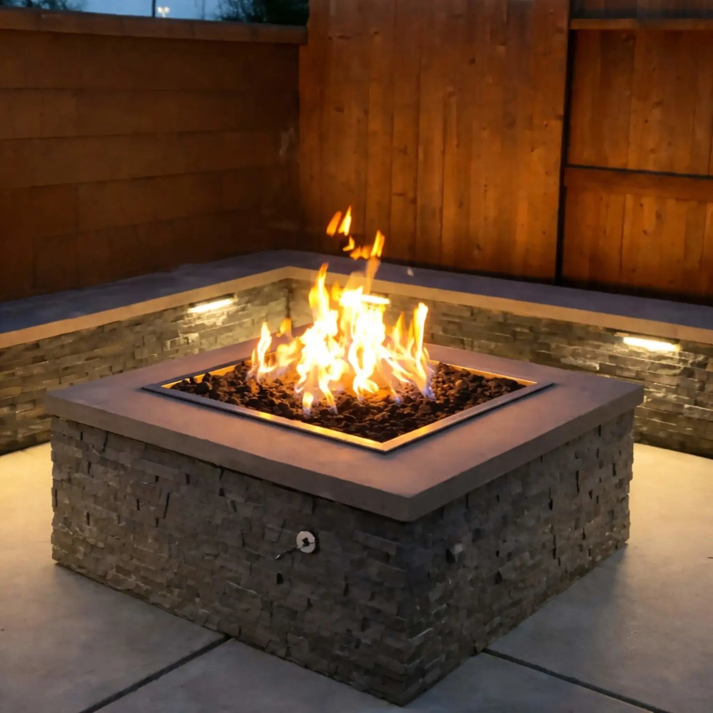 HPC Fire Inspired Hearth Products square drop-in electronic ignition on off hi lo low standard torpedo penta burner glowing at dusk in a modern backyard. The stone fire pit is filled with black lava rock and vibrant flames, surrounded by a wooden privacy wall with integrated lighting, creating a warm and inviting nighttime outdoor living space.