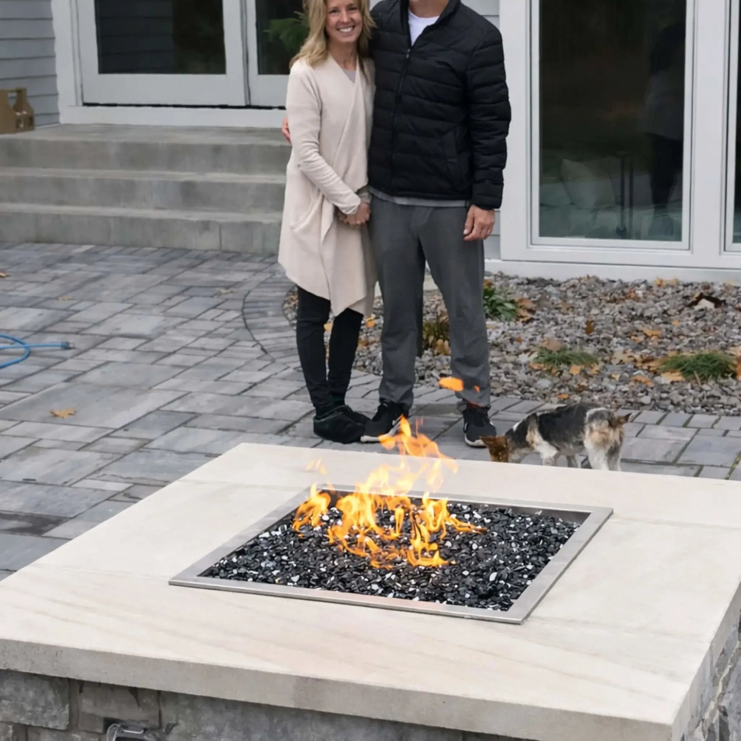 HPC Fire Inspired Hearth Products square drop-in electronic ignition on off hi lo low standard torpedo penta burner featured in a residential backyard fire table with black fire glass media. A smiling couple stands behind the fire pit with a small dog nearby, illustrating real-life use and scale of the fire feature in a casual outdoor patio setting.