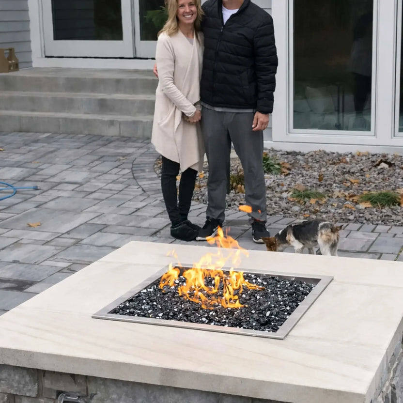 HPC Fire Inspired Hearth Products square drop-in electronic ignition on off hi lo low standard torpedo penta burner featured in a residential backyard fire table with black fire glass media. A smiling couple stands behind the fire pit with a small dog nearby, illustrating real-life use and scale of the fire feature in a casual outdoor patio setting.