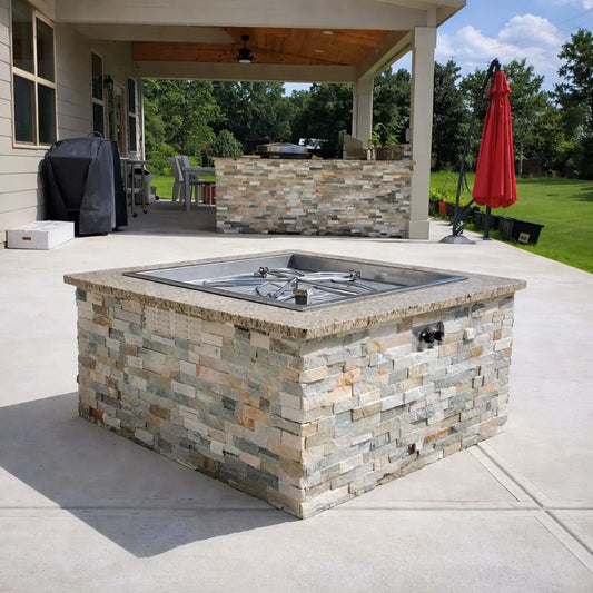 HPC Fire Inspired Hearth Products torpedo penta burner installed in a stone veneer fire pit on a covered patio, shown unlit to highlight the square drop-in pan and burner layout. Outdoor kitchen elements, grill area, and open yard emphasize a functional and stylish backyard setup.
