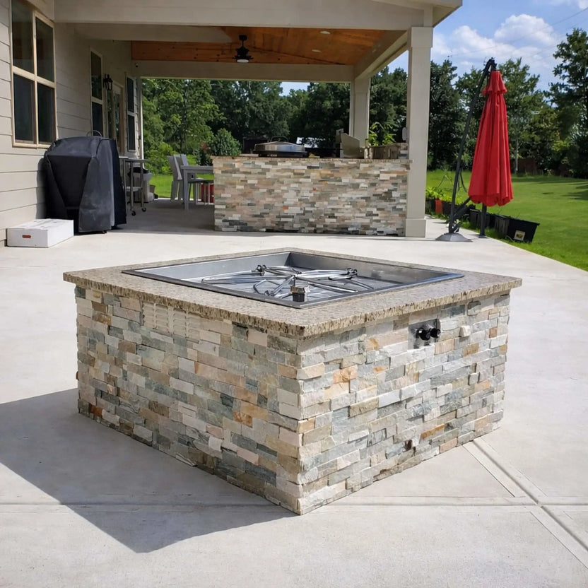 HPC Fire Inspired Hearth Products torpedo penta burner installed in a stone veneer fire pit on a covered patio, shown unlit to highlight the square drop-in pan and burner layout. Outdoor kitchen elements, grill area, and open yard emphasize a functional and stylish backyard setup.