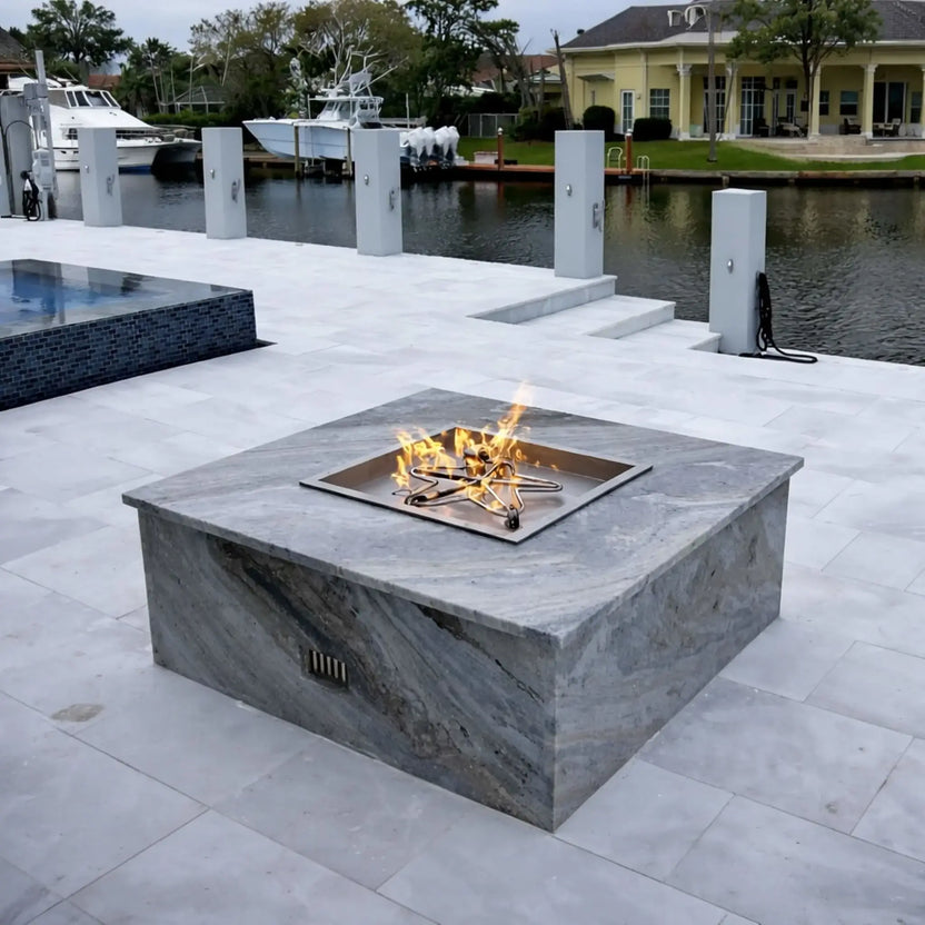 HPC Fire Inspired Hearth Products torpedo penta burner installed in a waterfront patio fire pit, with flames rising from a square drop-in pan. The background features docks, boats, and upscale homes, showcasing a premium outdoor lifestyle and modern fire feature integration.