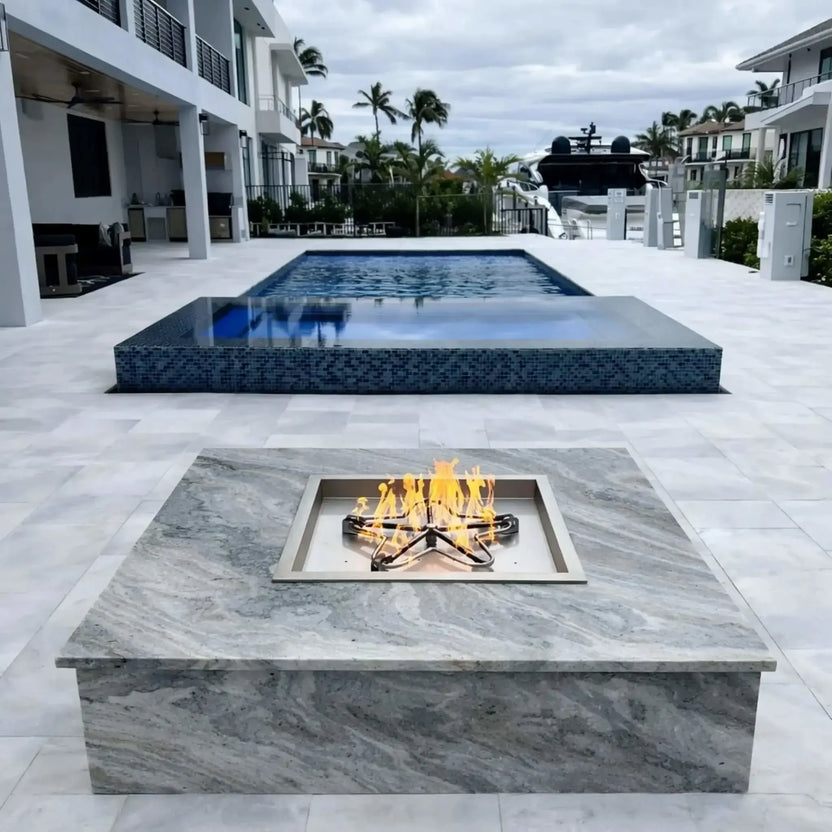 HPC Fire Inspired Hearth Products square drop-in match lit torpedo penta burner installed in a modern stone fire pit table beside a luxury pool. Flames rise evenly from the burner, showcasing a clean, contemporary outdoor living space with tiled pool, waterfront view, and upscale patio design ideal for entertaining.