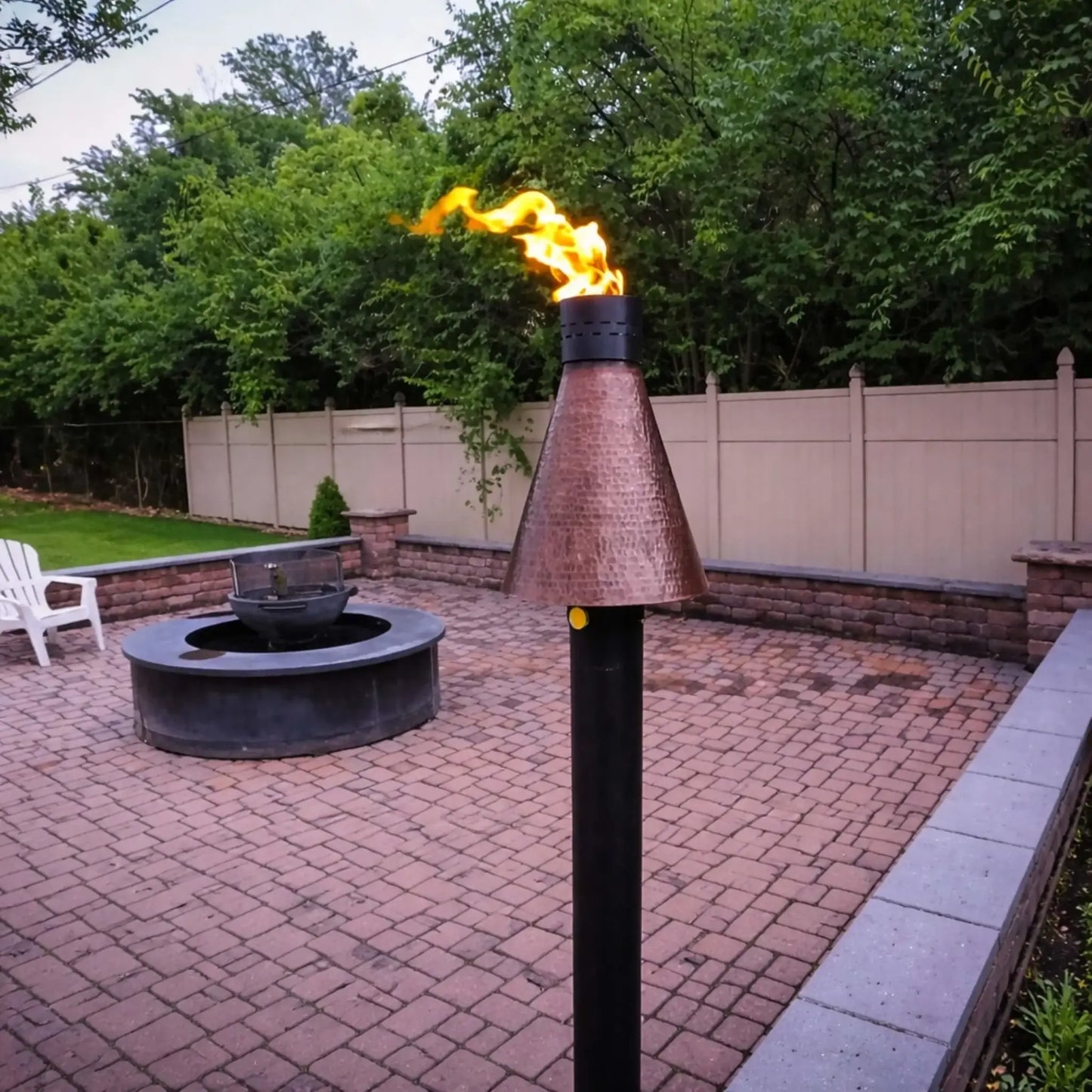 HPC TK Torch Copper Head and Post installed on a patio with paver flooring, actively burning with a bright flame near a circular fire pit, demonstrating how the hammered copper torch complements outdoor living spaces and fire feature layouts.
