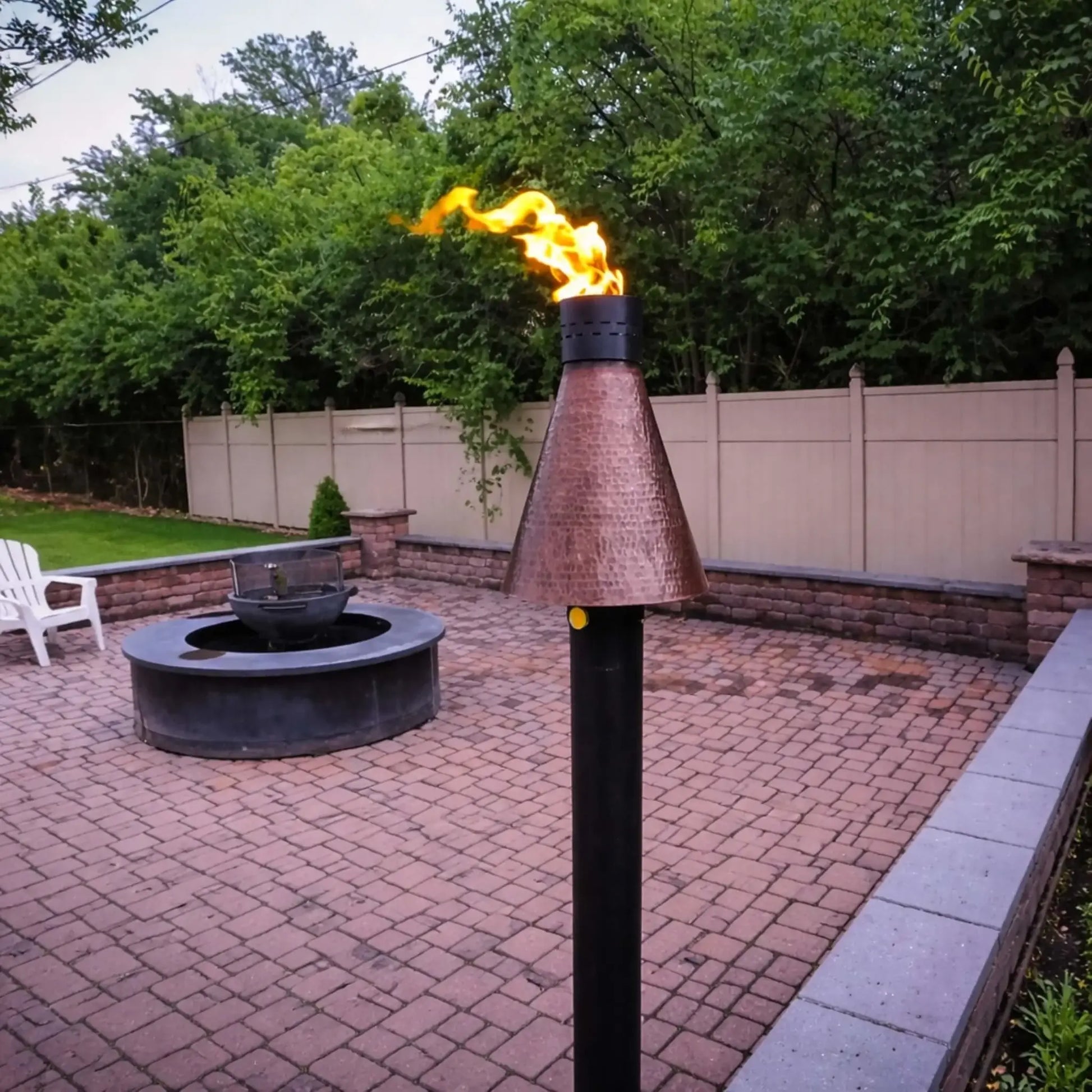 HPC TK Torch Copper Head and Post installed on a patio with paver flooring, actively burning with a bright flame near a circular fire pit, demonstrating how the hammered copper torch complements outdoor living spaces and fire feature layouts.