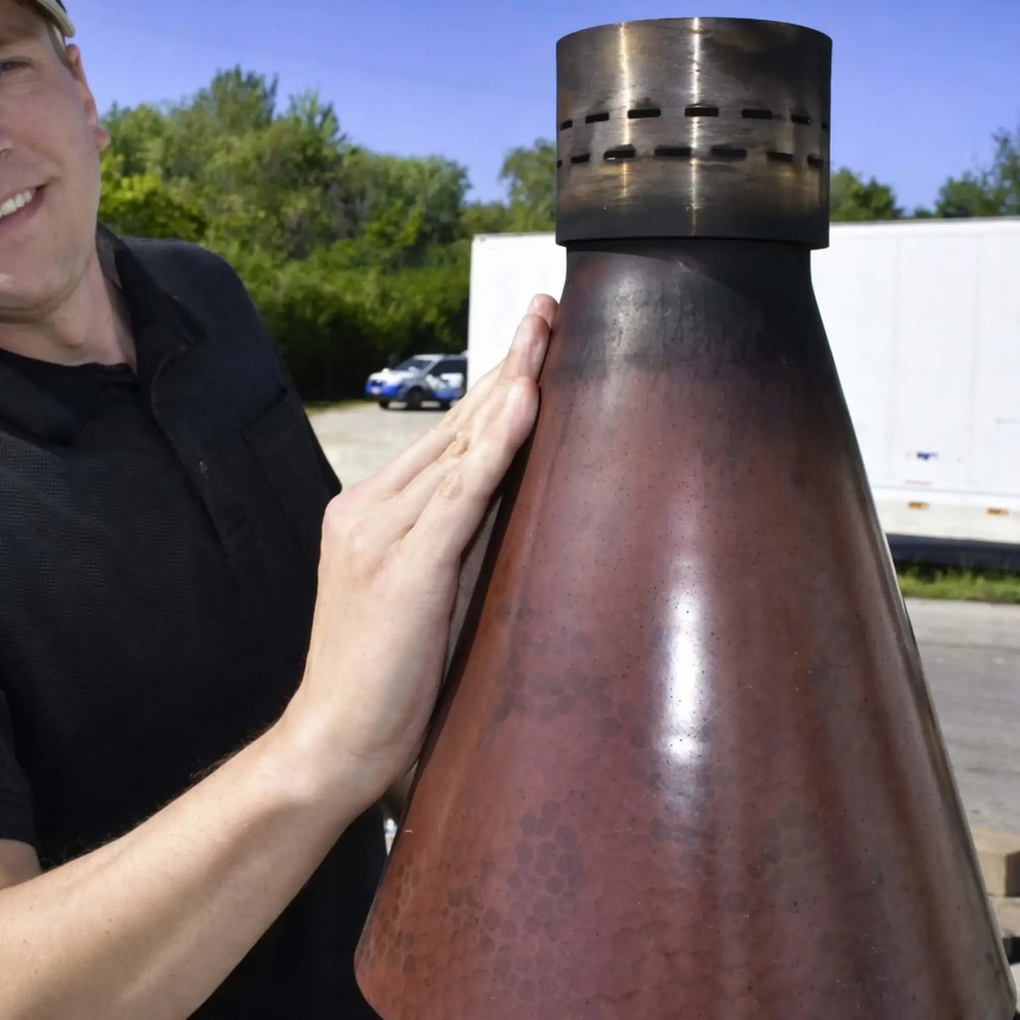 HPC TK Torch Copper Head and Post Lifestyle 10 showing a close-up of the copper torch head being handled outdoors, highlighting the smooth patina finish, vented top, and craftsmanship details against a natural outdoor background.