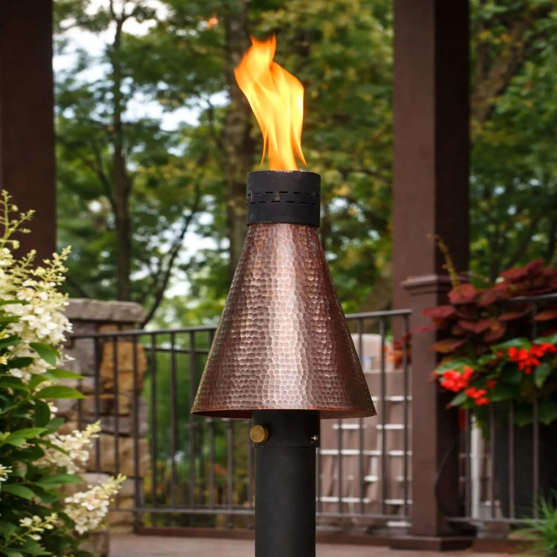 HPC TK Torch Copper Head and Post lit in a landscaped backyard, positioned near a deck with railing and flowering plants, highlighting the warm glow of the flame and the textured copper finish enhancing the outdoor ambiance.