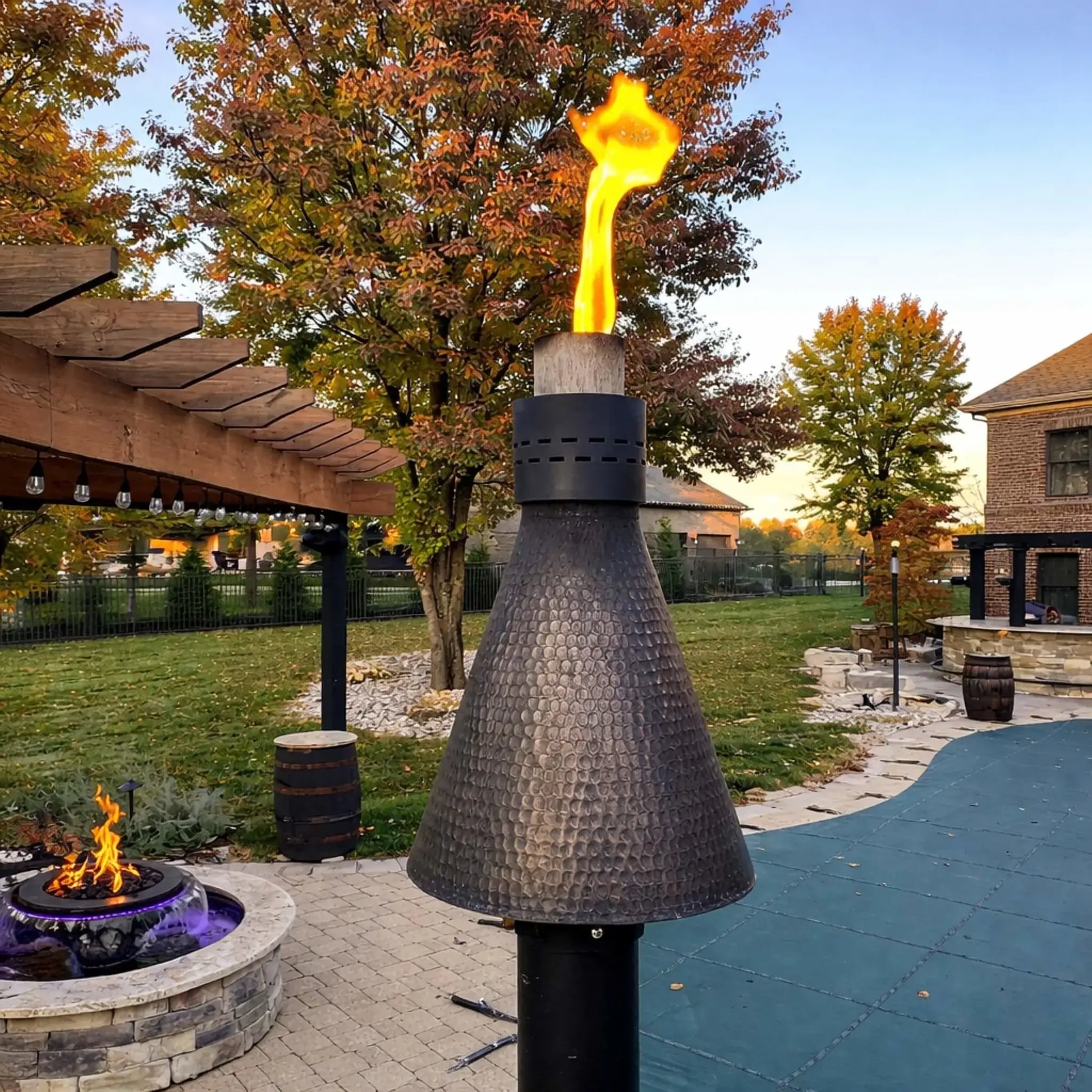 HPC Fire Inspired Hearth Products Controls TK Torch Copper Head and Post Lifestyle 4 showing a hammered copper torch burning with a tall flame beside a backyard pergola and circular fire pit, surrounded by autumn trees and paver patio, highlighting the torch’s warm glow and upscale outdoor living design.
