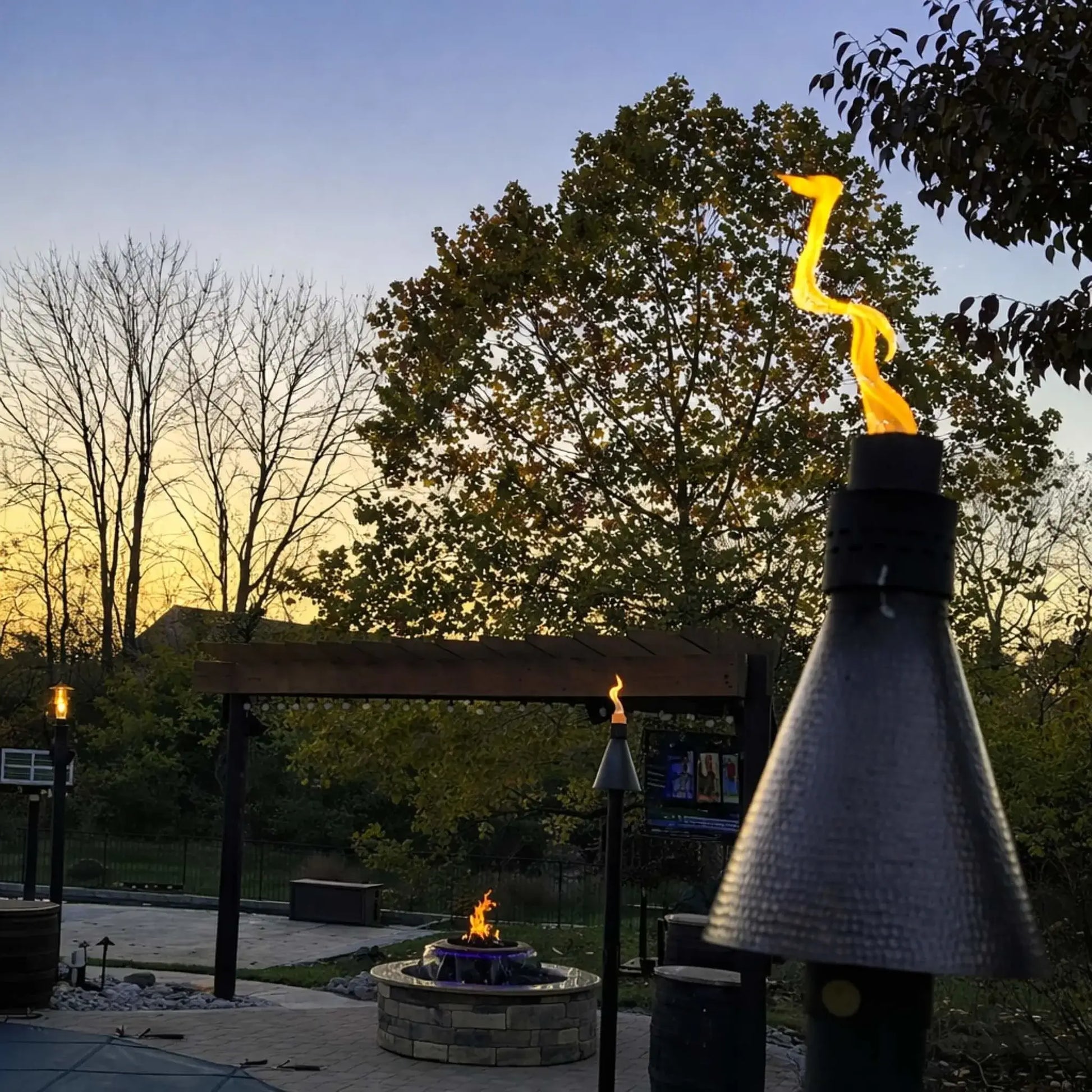 HPC TK Torch Copper Head and Post Lifestyle 5 captured at sunset with multiple torches lit around a patio, featuring a pergola, fire pit, and soft evening sky, showcasing how the copper torch enhances ambiance with natural flame lighting in a residential outdoor space.