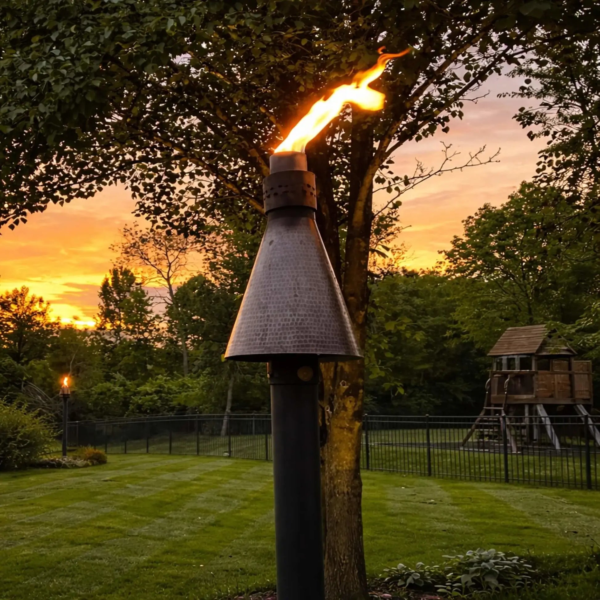 HPC TK Torch Copper Head and Post Lifestyle 6 displaying a single torch burning in a landscaped backyard at sunset, with vibrant orange sky and mature trees, emphasizing the hammered copper finish and dramatic flame presentation in a serene outdoor setting.